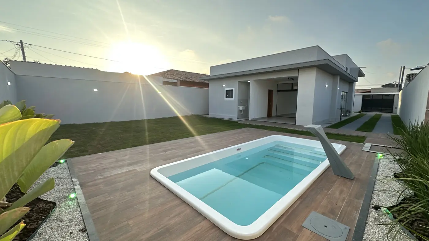 Piscina retangular com deck de madeira no quintal ao fim da tarde, com a casa iluminada ao fundo.