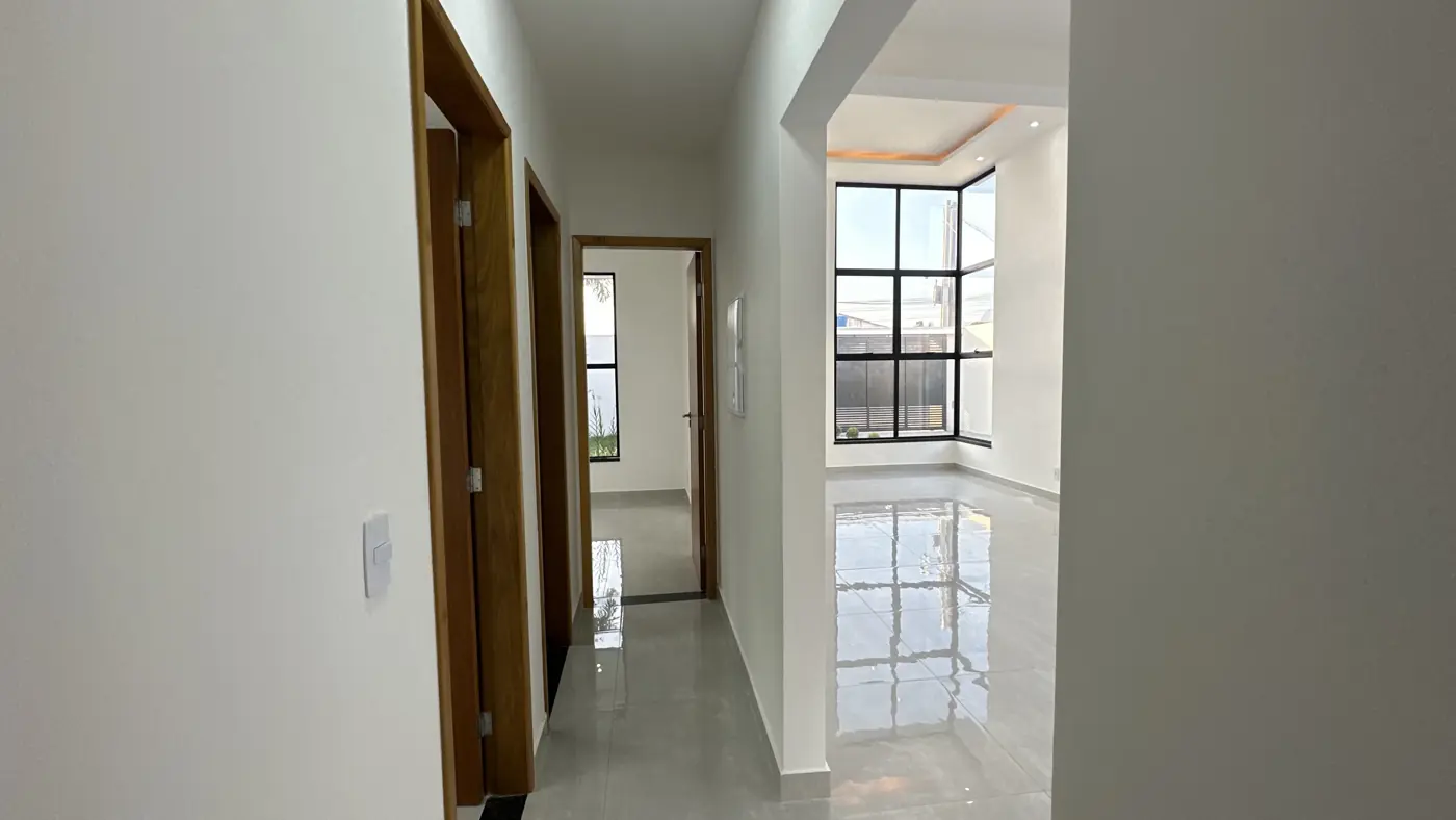 Corredor interno com portas de madeira e piso claro polido, com a sala iluminada ao fundo.