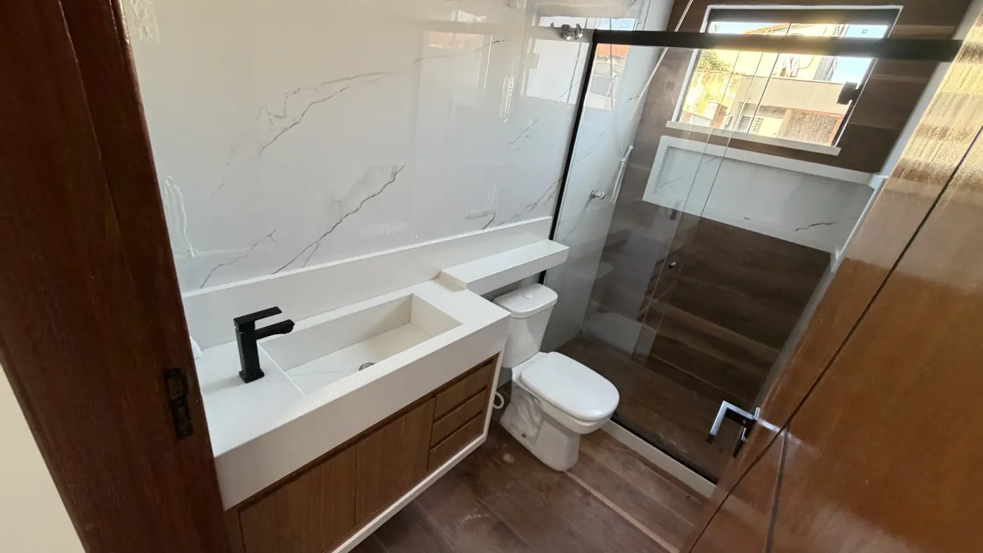 Banheiro com bancada branca esculpida, torneira preta, revestimento de mármore branco, armário de madeira e box de blindex.