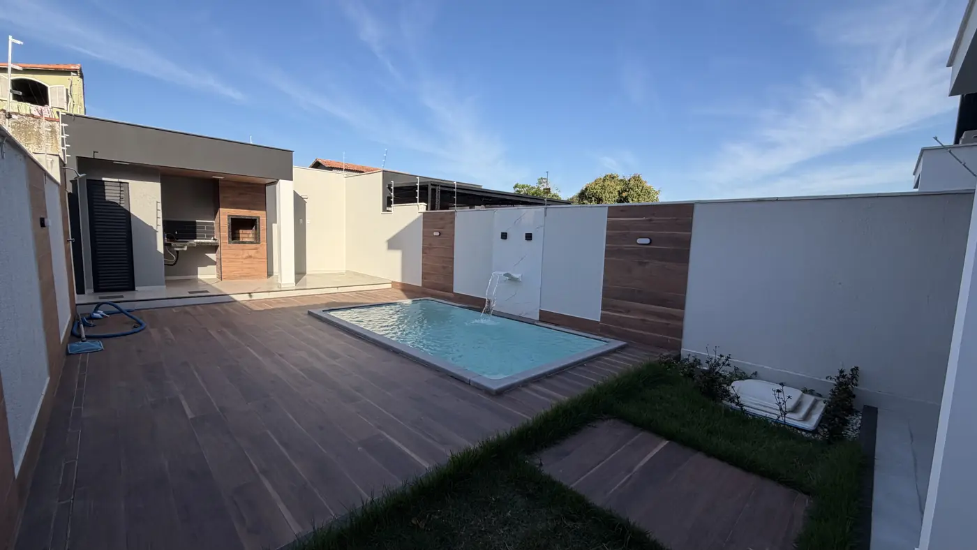 Quintal visto de outro ângulo: deck de madeira em volta da piscina, faixa de grama e a área gourmet coberta ao fundo.